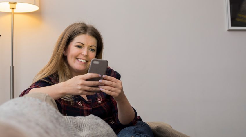 Woman sits on couch using her phone to pay rent