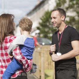 A male housing officer talking to a female customer holding a small child on the pavement next to houses 